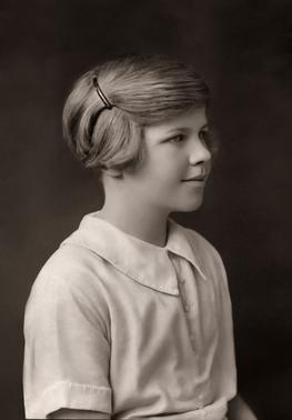 Head-and-shoulders black and white photograph of subject as a young girl. She wears a light-coloured blouse and faces right, looking out of the picture, with a slight smile. Her short hair is pulled back from her face and pinned up.