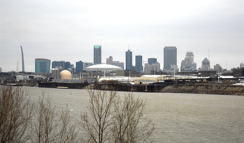 A large arch is in the center, across from a river. A clump of tall buildings is scattered behind it.