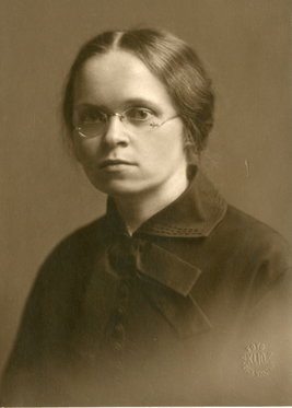 Sepia photograph of the torso and head of a woman wearing glasses in a 1920s coat