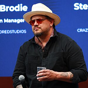 Sterlin Harjo wearing a black shirt, brown neckerchief, white Stetson hat, and red-shaded glasses, holding a plastic cup in one hand and a microphone in the other, standing onstage