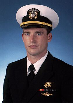 Color photograph of Michael Murphy, a U.S. Navy officer, wearing a military dress uniform. There is a blue background behind him and he is wearing a gold Navy Seal Trident, two blue and green striped ribbons, one red and yellow striped ribbon and gold parachute insignia wings below the ribbons.