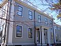 A gray two story house with white trim surrounding black-framed windows. The front door is sheltered by a small pillared porch, and there is a balustered railing around the roof.