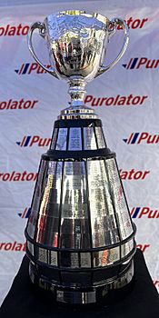 A trophy in the design of a silver cup affixed to a large, round wooden base.  The base has silver plates attached to it engraved with the names of previous winners.