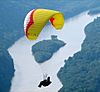 A person dangles below a red, yellow and white parasail, in the background is a lush forest with a large river and a tree-covered island.