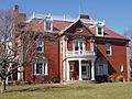 A brick two story house with richly detailed tan and white trim. A round portico with balcony above is in the center of the house, and the upper roof section has a low white railing surrounding it.