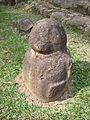 A small stone sculpture with a round featureless head and squat round body. The figure is seen from the front left and the left arm is visible, raised against the side of the figure's body. The arm descends from the shoulder and bends forwards at the elbow, running horizontally to the wrist, which bends sharply downwards, with the fingers marked by carved grooves. The sculpture has no legs and rests on a grassy area in front of some stone steps.