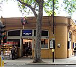 An orange building with an illuminated, blue sign reading "HOLLAND PARK STATION" in white letters and people walking in front all under a white sky