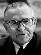 Arms wearing a dark jacket, white shirt, dark tie and thick spectacles.