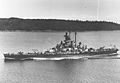 A battleship sails on calm waters with land in the background.