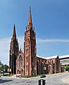 A church of ornate brown stone with two tall towers in front seen from across an intersection.