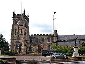 alt=A large medieval sandstone church, with tower, a clock face and stained glass windows. A statue of an angel of peace in the foreground