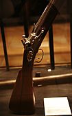 Well-polished stock, firing mechanism and lower barrel of a vertically-mounted musket. The musket is standing within a case, with an illegible white label at lower right.