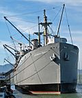 Photograph of the liberty ship SS "Jeremiah O'Brien" at dock, cranes bristling along its length. A banner on a nearby fence reads "Open", indicating its status as a museum ship.