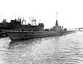 A long low-slung naval ship is entering a dock. Its deck is lined with sailors.