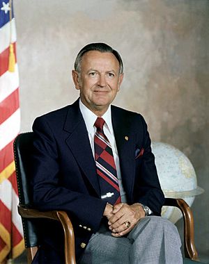 Formal portrait of a man in a jacket and tie seated in a chair in front of a U.S. flag.