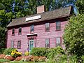 A red two story colonial house with wooden roof. Flowers have been planted on either side of the front door.