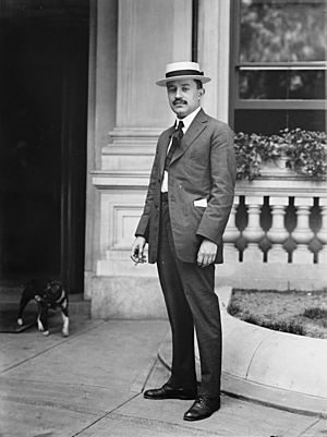 A black-and-white portrait of a formally&dressed young man with a short, black mustache wearing a light-colored hat, white shirt, a light-colored suit, a dark tie, and dark shoes. The man is outside a building from which a dog is coming out.