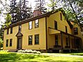 A two-story colonial-style house, painted yellow with brown trim. A porch with benches on it is off the right side of the house.