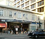 A light grey building with "M.R. ALDGATE STATION M.R." written in stone on the front face and a black car driving in the foreground