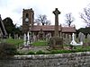 A church seen from the south; to the left is the tower with a round blue clock face, in the middle is the nave, and at a lower level to the right is the chancel. Immediately in the foreground is a wall behind which is a large monument surmounted by a Celtic cross; behind this are many gravestones