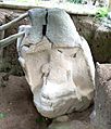 A stone sculpture sitting in an excavated trench. The monument is damaged with deep fracturing towards the top. It lists to the left and is braced with wooden poles. A niche is carved into the front of the monument and contains a badly eroded seated figure holding a featureless bundle in its arms. 