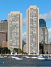 Distant view of two waterfront skyscrapers; both have Brutalist-style architecture, with concrete facades. Though very similar in appearance, the building on the right is taller and thinner. Several anchored sailboats are visible in the foreground.