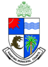 A quartered shield, blue and white. Top left has a red sun. Top right has blue waves. Bottom left has a crocodile. Bottom right has a coconut tree. Above, a green coat of arms of the Gambia. Below, a banner saying "KANIFING MUNICIPAL COUNCIL".