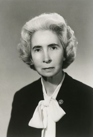 Black and white image of a woman with white hair wearing a black coat and white scarf tied in a bow.