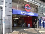 A grey-panelled building with a blue sign reading "FINSBURY PARK" in white letters and a woman with a brown coat standing in the doorway