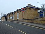 A brown-bricked building with a rectangular, dark blue sign reading "PINNER" in front of a road all under a cloudless, light blue sky