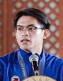 Carlos Yulo in blue tracksuit with medal speaking on a podium