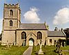 Stone building with square tower at left hand end. In the foreground either side of a path are gravestones in a grassy area.