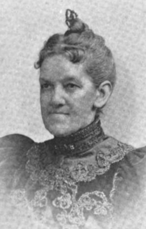 A white woman, hair dressed to the crown, wearing a high-collared dark dress with elaborate lace trim on the bodice, and puffed sleeves