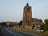 A curving road with buildings in the distance on the left and a church with a relatively large tower on the right. In front of the church are gravestones, a wall, and signs reading "Village Road" and "Long Lane"