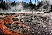 Blackened basin with orange streaks; steam is rising from it with fir trees in the background