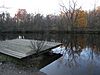 A wooden dock on the shore of a small lake in which autumnal trees are reflected