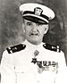 Head and shoulders of a white man with a thin, upturned mustache wearing a white peaked cap with black visor and a white jacket with dark shoulder boards, a row of medals on the left breast, and a star–shaped medal hanging from the neck.