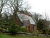 A small church seen from the southwest with a south porch. At the far end a saddleback-roofed tower is just visible. Around the church are trees and bushes and in the foreground is a wall