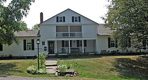 A large white house, built in a style similar to farmhouse styles. There is a large porch out front, with a large green yard.