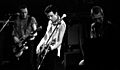 The rock band The Clash performing onstage. Three members are shown. All three have short hair. Two of the members are playing electric guitars.