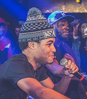 Jay Whiss wearing dark t-shirt and tuque with pompom, squatting among a group of people, holding microphone