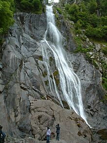 A large waterfall tumbling over grey igneous rock, with people below observing it.