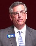 Brad Raffensperger wearing a grey suit with a name tag.