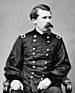 A white man with a Van Dyke beard and mustache sitting in a chair, wearing a baggy double-breasted military jacket.