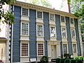 A three story colonial house with blue wood siding in front and brick walls on the sides.