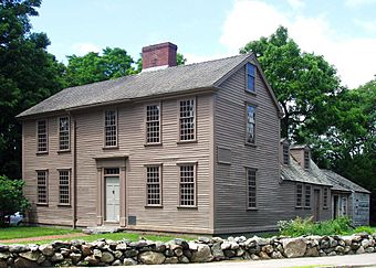 The main part of the home is a wooden, 2+1⁄2-story rectangular building with large windows, one central door, and a central chimney. A smaller wing extends back from the right side. There are large trees in the background and a low rock wall in the foreground.