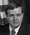 Black-and-white photo of a man in a suit and combed-over hair with the US flag behind him