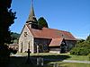 A church built in irregular stones with a red tiled roof seen from the southwest. On the west front is a round window and above this is a small spire. Protruding from the south wall is a red-tiled, timber-framed porch