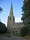 A tall spire is on the left and part of the body of the church is on the right. Steps lead to a porch and the whole is framed by trees