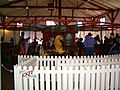 A wooden carousel with colorfully painted horses stands behind a white fence. There are parents and children on the carousel.